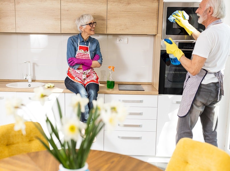 Elderly Caucasian man wiping down the oven exterior in the kitchen while his wife sits on the cabinets talking to him