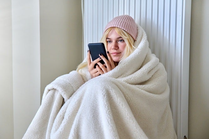 Young blonde Caucasian woman looking at her phone sitting against a radiator with a fleece blanket wrapped around her